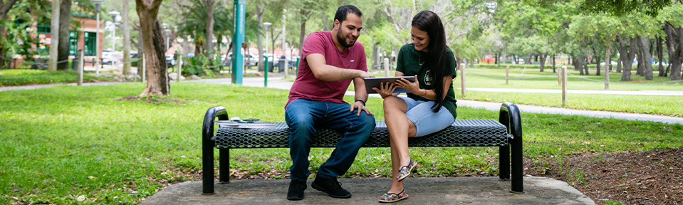Two ֱ students sitting on a bench and using a tablet.