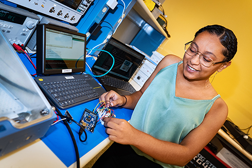 A student holds wires from an engineering kit next to a computer with coding on the screen.