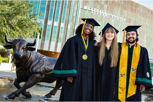 three students in full graduation regalia stand outside the marshall center, with a bull statue to their left