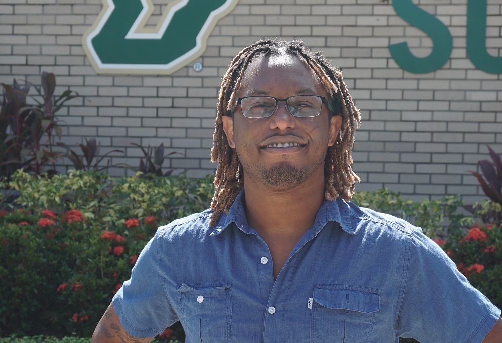 Dark-skinned male with dreadlocked hair and glasses standing in front of a ֱ sign wearing a demin shirt.