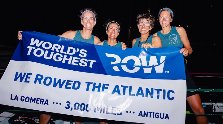 ֱ team, Salty Science, standing abroad their ship holding a banner proclaiming their successful finish of the World’s Toughest Row-Atlantic race